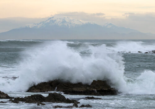 A wave crashes against rocks on the coast with a snow-capped mountain in the background and a moody yellow sunset.